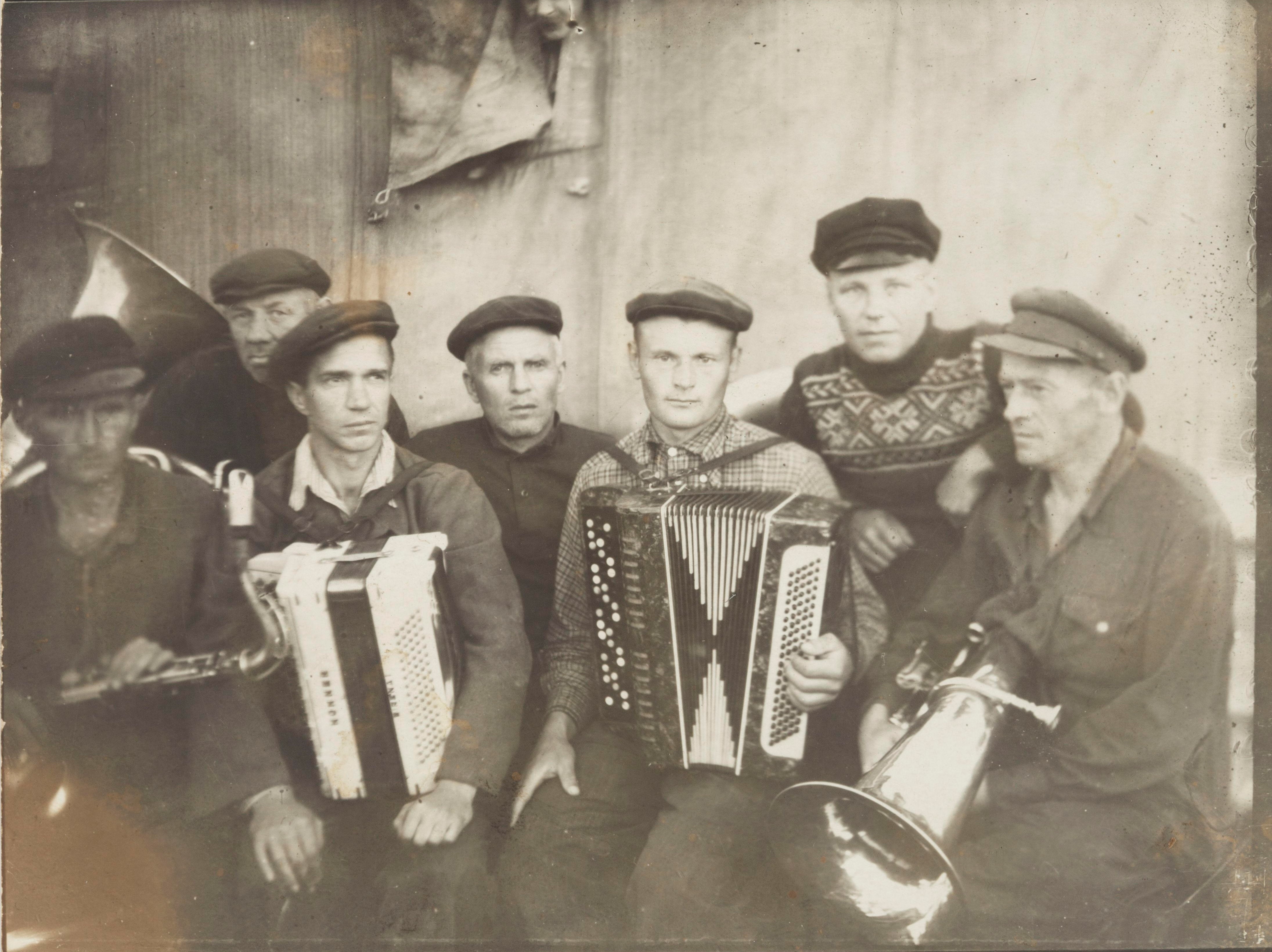 Yhteiskuva orkesterista Nadvoitsan vankileirillä. Suomalainen Vilho Saarinen on kuvassa vasemmalla tenorisaksofoni sylissään. Leirivangeista koostuvat orkesteri oli varsin kansainvälinen: muut soittajat olivat kansallisuudeltaan liettualaisia, karjalaisia, venäläisiä, virolaisia ja latvialaisia. Kuva on vuodelta 1954, viimeiseltä kesältä ennen Saarisen leiriltä vapautumista. SKS KRA, Hannele Mikkelän kokoelma. CC BY-NC-ND 4.0